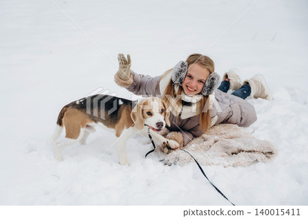 Young girl with long blonde hair wearing earmuffs and gloves is playing with a beagle dog in the snow, lying on a blanket during winter fun in a snowy landscape Young girl with long blonde hair wearing earmuffs and gloves is playing with a beagle dog in the snow, lying on a blanket during winter fun in a snowy landscape 140015411