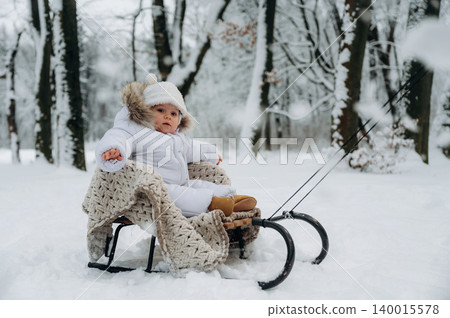 Baby in white winter outfit sits on sled covered with cozy blanket, surrounded by snowy trees, enjoying a cold day outdoors, capturing the essence of winter fun and joy Baby in white winter outfit sits on sled covered with cozy blanket, surrounded by snowy trees, enjoying a cold day outdoors, capturing the essence of winter fun and joy 140015578