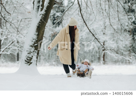 Woman in cozy winter attire joyfully pulls a sled with a child through a snowy landscape, surrounded by trees, capturing the essence of winter fun and family bonding 140015606