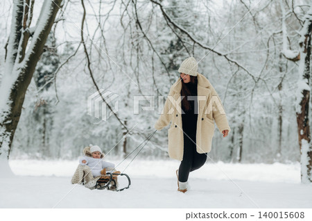 Woman in cozy winter coat pulls child on sled through snowy landscape, surrounded by trees, creating a joyful winter scene of family fun and outdoor adventure 140015608