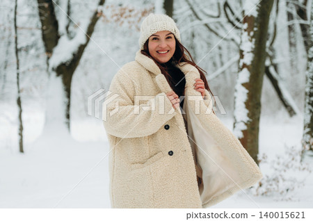 Smiling woman in cozy cream coat and knitted hat stands in snowy forest, surrounded by trees, showcasing winter fashion and joyful atmosphere in nature Smiling woman in cozy cream coat and knitted hat stands in snowy forest, surrounded by trees, showcasing winter fashion and joyful atmosphere in nature 140015621