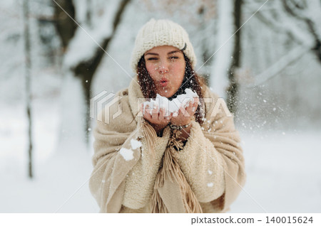 Young woman with long hair, wearing a cozy winter outfit, joyfully blowing snowflakes from her hands in a serene snowy forest, capturing the essence of winter fun and joy 140015624