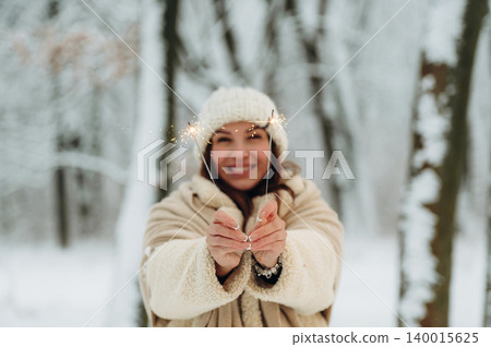 Smiling woman with dark hair wearing cozy winter clothing is holding sparklers in a snowy forest, celebrating joy and warmth during the holiday season with festive spirit Smiling woman with dark hair wearing cozy winter clothing is holding sparklers in a snowy forest, celebrating joy and warmth during the holiday season with festive spirit 140015625
