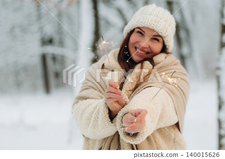 Smiling woman with long hair, wearing cozy winter attire, holding sparklers in a snowy forest, celebrating joy and warmth during festive season with copy space Smiling woman with long hair, wearing cozy winter attire, holding sparklers in a snowy forest, celebrating joy and warmth during festive season with copy space 140015626
