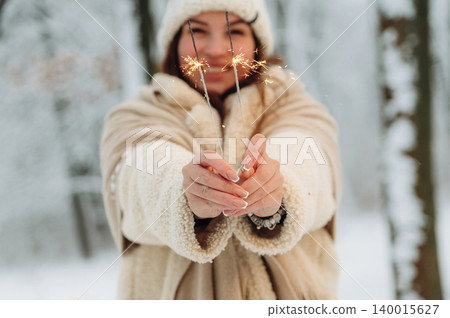 Young woman with brown hair, wearing cozy winter attire, joyfully holding sparklers in a snowy forest, celebrating the festive season with warmth and cheer 140015627