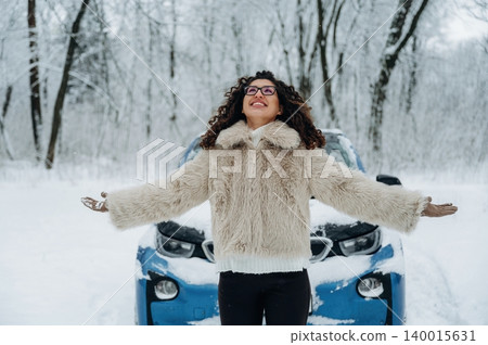 Young woman with curly hair wearing a fur coat stands joyfully in a snowy landscape with a blue car in the background surrounded by frosty trees and winter scenery 140015631