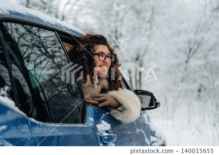Woman with curly hair wearing a fur coat leans out of a blue car window, smiling in a snowy landscape with trees covered in white during winter season Woman with curly hair wearing a fur coat leans out of a blue car window, smiling in a snowy landscape with trees covered in white during winter season 140015655