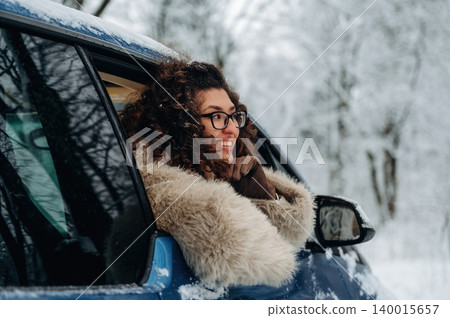 Woman with curly hair wearing a fur coat leans out of a blue car window, smiling in a snowy landscape with trees covered in white snow during winter season Woman with curly hair wearing a fur coat leans out of a blue car window, smiling in a snowy landscape with trees covered in white snow during winter season 140015657