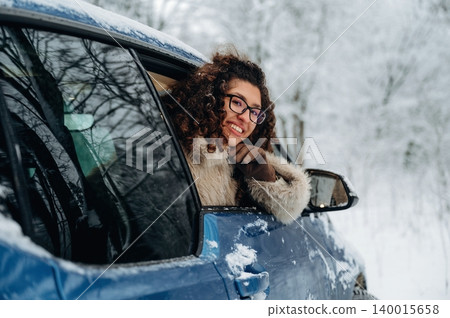 Young woman with curly hair and glasses is smiling from the window of a blue car parked in a snowy landscape with trees covered in frost during winter season Young woman with curly hair and glasses is smiling from the window of a blue car parked in a snowy landscape with trees covered in frost during winter season 140015658