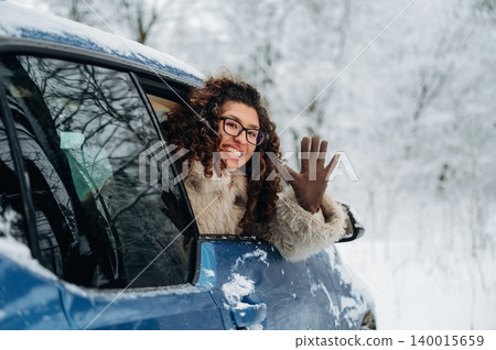 Woman with curly hair wearing a fur coat is waving from the window of a blue car in a snowy landscape with trees covered in white snow during winter 140015659