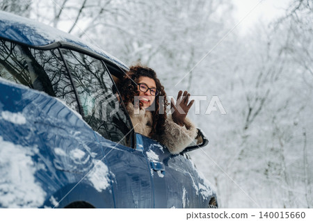 Young woman with curly hair wearing glasses is smiling and waving from the window of a blue car parked in a snowy landscape with trees in the background during winter 140015660