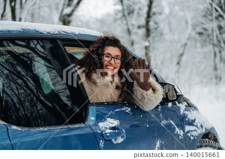 Young woman with curly hair wearing a fur coat is waving from the window of a blue car in a snowy landscape surrounded by trees during winter season Young woman with curly hair wearing a fur coat is waving from the window of a blue car in a snowy landscape surrounded by trees during winter season 140015661