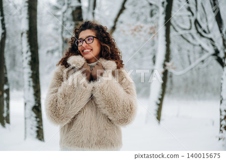 Young woman with curly hair wearing a fluffy beige coat smiles in a snowy forest, surrounded by tall trees and a winter landscape with soft white snow on the ground Young woman with curly hair wearing a fluffy beige coat smiles in a snowy forest, surrounded by tall trees and a winter landscape with soft white snow on the ground 140015675