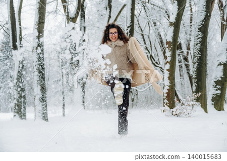 Young woman with curly hair wearing a cozy sweater is joyfully running through a snowy forest, kicking up snowflakes in a winter wonderland scene with trees in the background Young woman with curly hair wearing a cozy sweater is joyfully running through a snowy forest, kicking up snowflakes in a winter wonderland scene with trees in the background 140015683