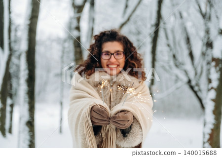 Woman with curly hair, wearing a cozy shawl, joyfully holding sparklers in a snowy forest, celebrating winter festivities with a cheerful expression and magical ambiance 140015688