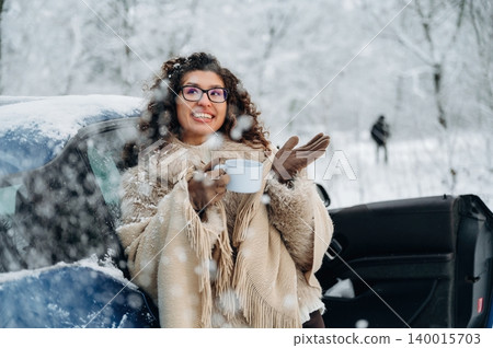 Woman with curly hair wearing a cozy sweater enjoys a warm drink while standing beside a car in a snowy landscape, capturing the essence of winter joy and comfort Woman with curly hair wearing a cozy sweater enjoys a warm drink while standing beside a car in a snowy landscape, capturing the essence of winter joy and comfort 140015703