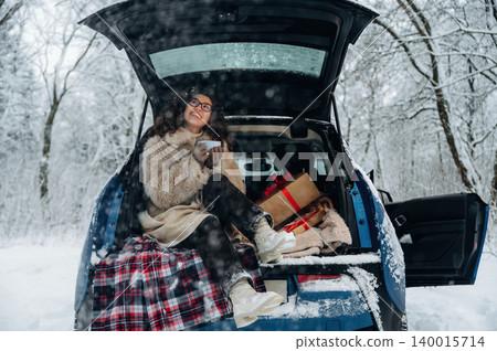 Young woman in cozy winter attire sits in car trunk, enjoying hot beverage surrounded by gifts, snow-covered trees creating a serene holiday atmosphere with copy space Young woman in cozy winter attire sits in car trunk, enjoying hot beverage surrounded by gifts, snow-covered trees creating a serene holiday atmosphere with copy space 140015714