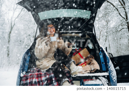 Woman enjoying a warm beverage while sitting in the back of a car, surrounded by snow-covered trees and holiday gifts, creating a cozy winter atmosphere with joy 140015715