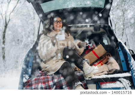 Woman in winter attire enjoys a warm drink while sitting in the back of a car, surrounded by festive gifts and a snowy landscape, creating a cozy holiday atmosphere Woman in winter attire enjoys a warm drink while sitting in the back of a car, surrounded by festive gifts and a snowy landscape, creating a cozy holiday atmosphere 140015716