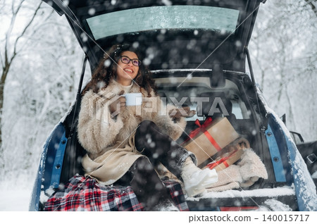 Woman in cozy winter attire enjoys a warm drink while sitting in the trunk of a car surrounded by snow-covered trees and festive gifts, embodying holiday cheer and relaxation Woman in cozy winter attire enjoys a warm drink while sitting in the trunk of a car surrounded by snow-covered trees and festive gifts, embodying holiday cheer and relaxation 140015717