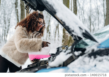 African American woman in a cozy winter coat is adding fluid to a car engine in a snowy forest, showcasing automotive maintenance in cold weather conditions African American woman in a cozy winter coat is adding fluid to a car engine in a snowy forest, showcasing automotive maintenance in cold weather conditions 140015718