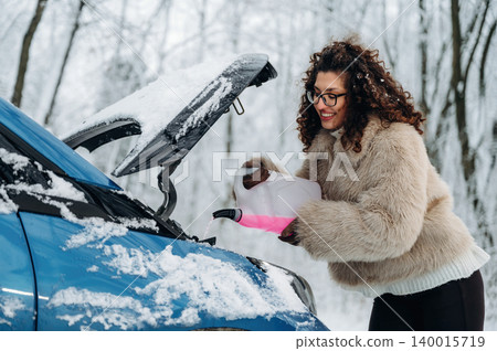 Woman with curly hair in a cozy fur coat is pouring liquid into the engine of a blue car in a snowy landscape, showcasing winter vehicle maintenance and care Woman with curly hair in a cozy fur coat is pouring liquid into the engine of a blue car in a snowy landscape, showcasing winter vehicle maintenance and care 140015719