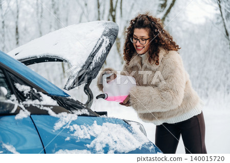 Woman with curly hair, wearing a cozy fur coat, is pouring fluid into the engine of a blue car in a snowy landscape, showcasing winter car maintenance and care Woman with curly hair, wearing a cozy fur coat, is pouring fluid into the engine of a blue car in a snowy landscape, showcasing winter car maintenance and care 140015720