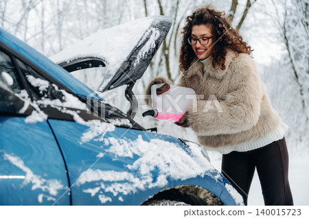 Woman with curly hair in a cozy fur jacket is pouring fluid into the engine of a blue car covered in snow, showcasing winter car maintenance and care Woman with curly hair in a cozy fur jacket is pouring fluid into the engine of a blue car covered in snow, showcasing winter car maintenance and care 140015723