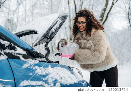 Woman in a cozy fur coat is pouring liquid into the engine of a blue car in a snowy landscape, showcasing winter vehicle maintenance and care in cold weather Woman in a cozy fur coat is pouring liquid into the engine of a blue car in a snowy landscape, showcasing winter vehicle maintenance and care in cold weather 140015724