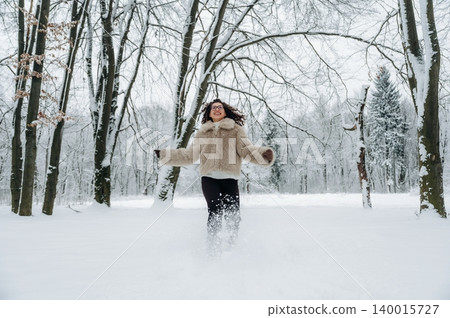 Woman in cozy fur coat joyfully running through deep snow in a winter forest, surrounded by tall trees and a serene white landscape, capturing the essence of winter fun 140015727