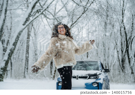Young woman with curly hair wearing a fur coat is joyfully standing in the snow beside a blue electric car in a winter forest setting with snow-covered trees Young woman with curly hair wearing a fur coat is joyfully standing in the snow beside a blue electric car in a winter forest setting with snow-covered trees 140015756
