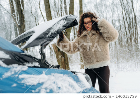Woman with curly hair in a fluffy coat is surprised while inspecting the engine of a blue car in a snowy forest during winter weather conditions Woman with curly hair in a fluffy coat is surprised while inspecting the engine of a blue car in a snowy forest during winter weather conditions 140015759