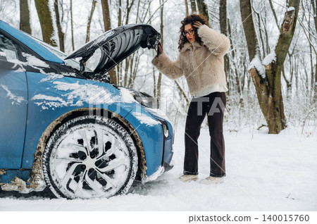 Female driver in a winter setting is inspecting her blue car with the hood open, surrounded by snow-covered trees and a snowy landscape during a cold day Female driver in a winter setting is inspecting her blue car with the hood open, surrounded by snow-covered trees and a snowy landscape during a cold day 140015760