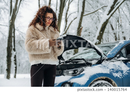 Young woman with curly hair wearing a fluffy coat is standing beside a blue car with an open hood in a snowy forest while checking her phone for assistance 140015764