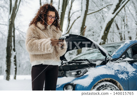 Young woman with curly hair wearing a fur coat is standing beside a blue car with an open hood in a snowy forest, checking her phone for assistance during a winter breakdown 140015765