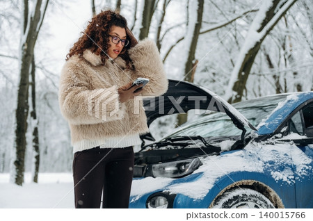 Young woman with curly hair in a fluffy coat is standing beside a blue car with an open hood in a snowy forest while using a smartphone for assistance Young woman with curly hair in a fluffy coat is standing beside a blue car with an open hood in a snowy forest while using a smartphone for assistance 140015766