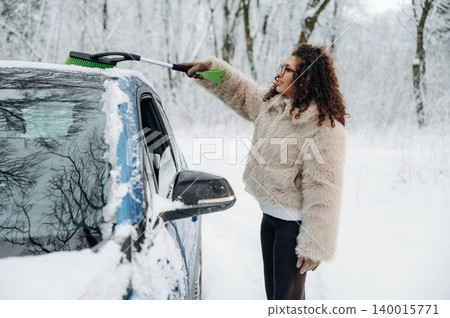 Woman with curly hair in a fur coat is removing snow from a blue car using a green snow brush in a snowy landscape with trees in the background during winter Woman with curly hair in a fur coat is removing snow from a blue car using a green snow brush in a snowy landscape with trees in the background during winter 140015771