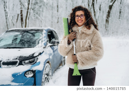 Young woman with curly hair wearing a fluffy coat is holding a snow shovel beside a blue car covered in snow in a winter forest setting with trees in the background Young woman with curly hair wearing a fluffy coat is holding a snow shovel beside a blue car covered in snow in a winter forest setting with trees in the background 140015785
