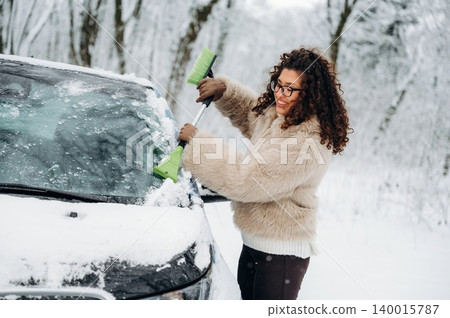 Woman with curly hair wearing a fur coat is clearing snow from a car windshield using a green snow brush in a snowy outdoor setting surrounded by trees Woman with curly hair wearing a fur coat is clearing snow from a car windshield using a green snow brush in a snowy outdoor setting surrounded by trees 140015787