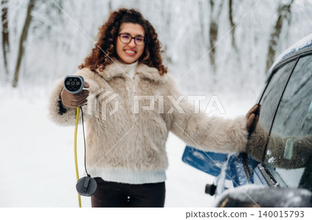 Woman with curly hair wearing a fluffy coat is holding an electric car charging cable near a vehicle in a snowy outdoor setting with trees in the background Woman with curly hair wearing a fluffy coat is holding an electric car charging cable near a vehicle in a snowy outdoor setting with trees in the background 140015793
