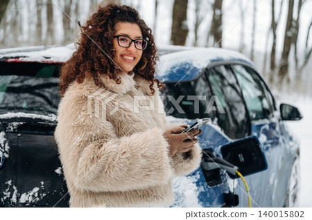 Young woman with curly hair wearing a fluffy coat is smiling while holding a smartphone near an electric car charging in a snowy outdoor setting with trees in the background Young woman with curly hair wearing a fluffy coat is smiling while holding a smartphone near an electric car charging in a snowy outdoor setting with trees in the background 140015802