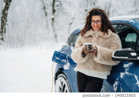 Woman with curly hair in a fur coat is standing beside a blue electric car in a snowy landscape while using a smartphone during winter charging Woman with curly hair in a fur coat is standing beside a blue electric car in a snowy landscape while using a smartphone during winter charging 140015803