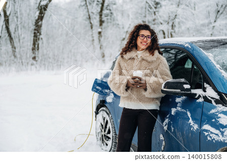 Young woman with curly hair wearing a fur coat is standing beside an electric car charging in a snowy landscape, holding a warm drink during winter Young woman with curly hair wearing a fur coat is standing beside an electric car charging in a snowy landscape, holding a warm drink during winter 140015808