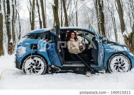 Electric car charging in snowy forest with a woman seated inside, wearing a cozy fur coat, holding a coffee cup, showcasing winter outdoor lifestyle and eco-friendly transportation Electric car charging in snowy forest with a woman seated inside, wearing a cozy fur coat, holding a coffee cup, showcasing winter outdoor lifestyle and eco-friendly transportation 140015811