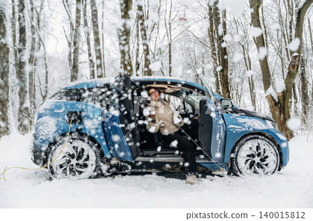 Electric car parked in snowy forest with charging cable connected, person relaxing inside vehicle, snow-covered trees creating a serene winter landscape with copy space Electric car parked in snowy forest with charging cable connected, person relaxing inside vehicle, snow-covered trees creating a serene winter landscape with copy space 140015812