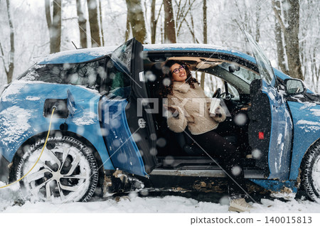Electric car parked in snowy forest with a woman enjoying a warm drink, charging cable connected, surrounded by snow-covered trees and a winter landscape with copy space Electric car parked in snowy forest with a woman enjoying a warm drink, charging cable connected, surrounded by snow-covered trees and a winter landscape with copy space 140015813