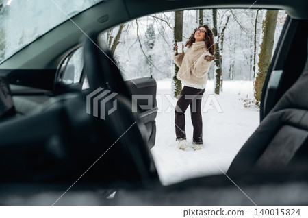 Young woman in a cozy white fur jacket stands outside a car in a snowy forest, surrounded by trees and winter scenery with snow-covered ground and a serene atmosphere Young woman in a cozy white fur jacket stands outside a car in a snowy forest, surrounded by trees and winter scenery with snow-covered ground and a serene atmosphere 140015824
