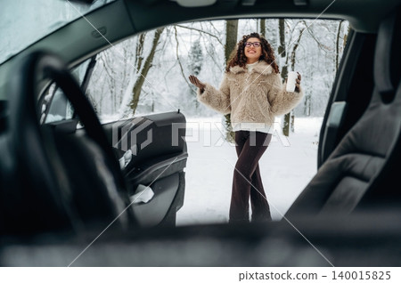 Young woman with curly hair in a cozy fur jacket stands by an open car door in a snowy landscape, holding a cup and enjoying the winter scenery with trees in the background Young woman with curly hair in a cozy fur jacket stands by an open car door in a snowy landscape, holding a cup and enjoying the winter scenery with trees in the background 140015825