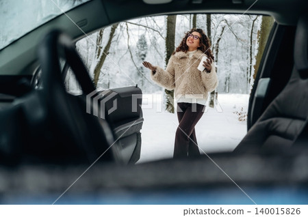 Young woman with curly hair wearing a fur coat stands outside a car in a snowy landscape, trees covered in snow visible in the background during winter season Young woman with curly hair wearing a fur coat stands outside a car in a snowy landscape, trees covered in snow visible in the background during winter season 140015826