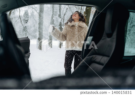 Woman with curly hair wearing a fur coat is enjoying snowfall outside a car in a winter landscape with trees and snow-covered ground visible in the background Woman with curly hair wearing a fur coat is enjoying snowfall outside a car in a winter landscape with trees and snow-covered ground visible in the background 140015831